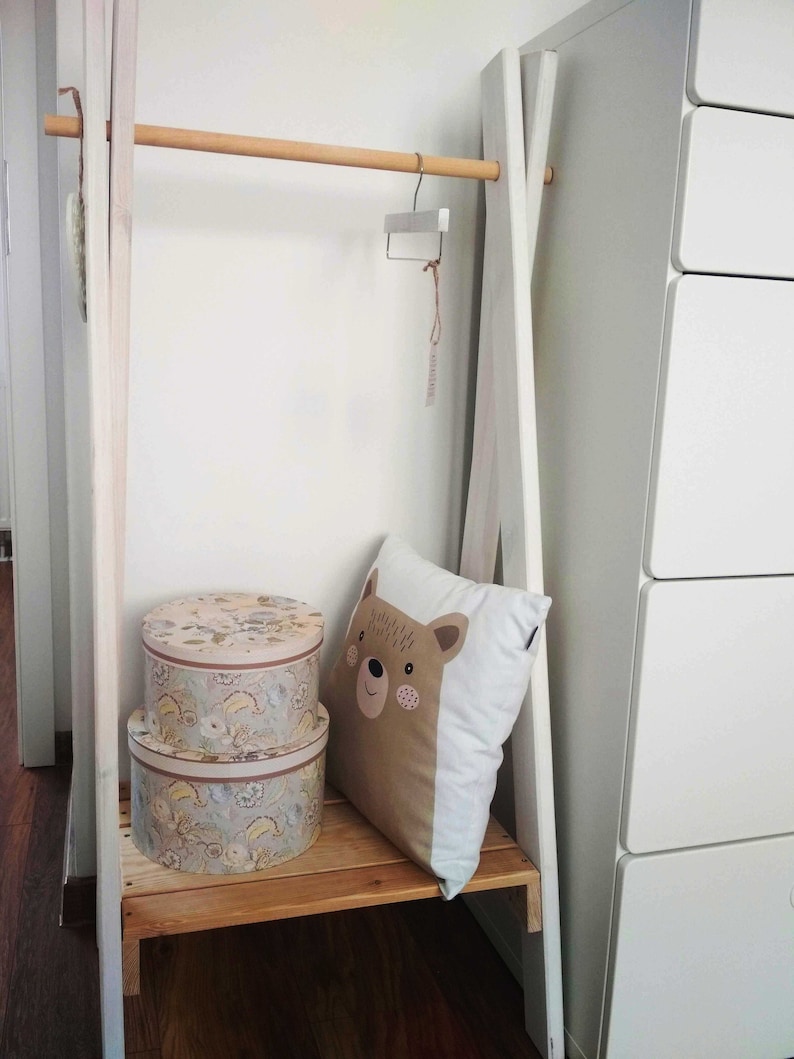 May include: White wooden clothing rack with a wooden bar, two round storage boxes with floral patterns, and a white pillow with a brown bear face design.
