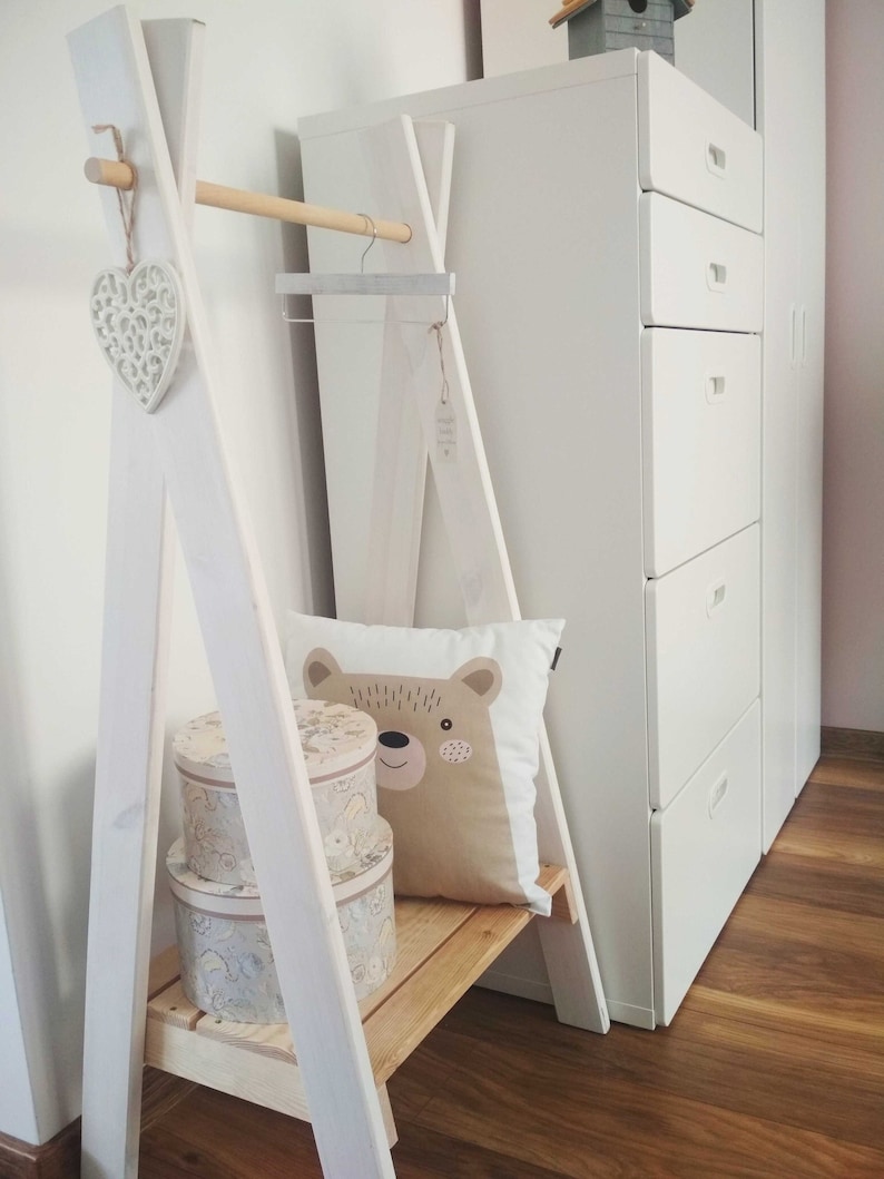 May include: White wooden A-frame clothing rack with a wooden dowel rod, a heart-shaped ornament, a brown bear pillow, and two round storage boxes with floral patterns. The rack has a wooden shelf and is sitting on a wooden floor.