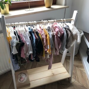 May include: White wooden clothing rack with a shelf. The rack is filled with a variety of colorful children's clothing on hangers.