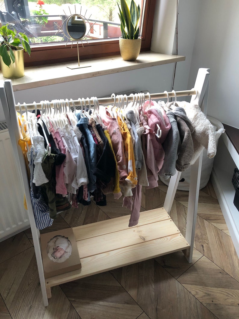May include: White wooden clothing rack with a shelf. The rack is filled with a variety of colorful children's clothing on hangers.