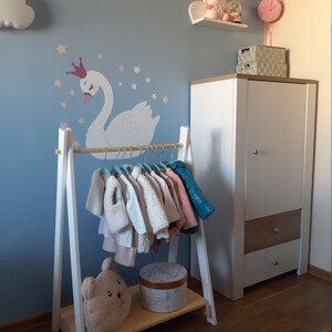 May include: White wooden clothes rack with a shelf, featuring a cute stuffed bear and a round box. The rack is decorated with a white swan and stars on a light blue wall.