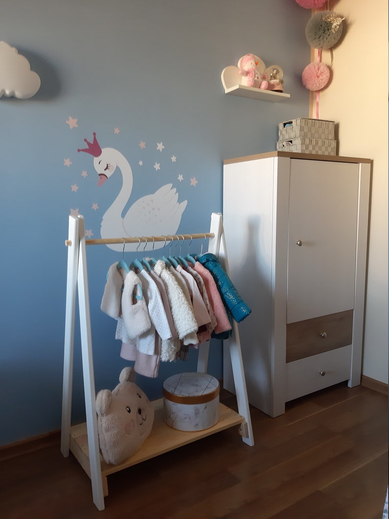 May include: White wooden clothes rack with a shelf, featuring a cute stuffed bear and a round box. The rack is decorated with a white swan and stars on a light blue wall.