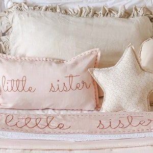 Puede incluir: Una cabecera de cama de metal blanco con una almohada con volantes blancos y beige, una almohada rosa con el texto "little sister" bordado en hilo de cobre y una almohada en forma de estrella beige con estampado floral.