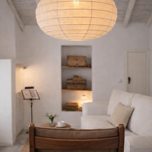 Puede incluir: Una lámpara de papel redonda de color crema cuelga del techo de una sala de estar. La lámpara está iluminada desde dentro, proyectando un brillo cálido. La habitación cuenta con un sofá blanco, una silla de madera y una mesa pequeña.