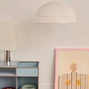 May include: A white paper dome pendant light hangs above a light blue metal modular shelving unit. On the shelf sits a lamp with a white shade, a dark red scalloped bowl, and a light green ceramic bowl. A painting of a person with stars is nearby.