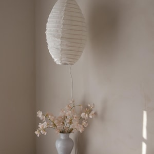 May include: A white paper lantern hangs from the ceiling, casting soft light. Below, a white vase holds delicate pink flowers, placed on a small table with a perfume bottle and a decorative plate. The scene evokes a sense of calm.