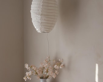 May include: A white paper lantern hangs from the ceiling, casting soft light. Below, a white vase holds delicate pink flowers, placed on a small table with a perfume bottle and a decorative plate. The scene evokes a sense of calm.