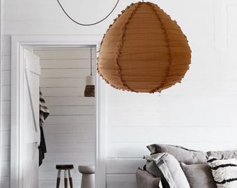 May include: A teardrop-shaped pendant light fixture with a textured, rust-colored fabric shade featuring vertical seams with frayed edges. The lamp hangs from a thin, dark cord that extends upward and across the ceiling, providing overhead illumination.