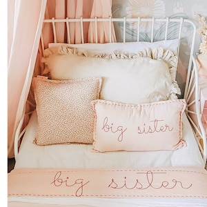 May include: A white metal bed frame with a pink and white bedding set. The bedding has the words "big sister" embroidered on it in red thread.