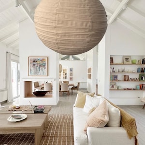 May include: A large, spherical, beige paper lantern pendant light fixture with a ribbed texture, suspended from a high, white, vaulted ceiling in a bright, open-concept living space with white walls and wooden furniture.