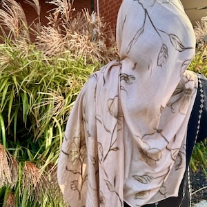 May include: A light pink scarf with a floral pattern in brown. The scarf is draped over a person wearing a black garment. The background features green and brown plants and a brick building.
