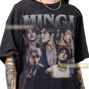 Peut inclure: T-shirt gris foncé avec un collage de cinq personnes et le nom "MINGI" en grandes lettres argentées. L'image a un aspect vintage et vieilli. Le t-shirt a des manches courtes et un col rond.
