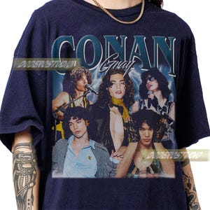 May include: Navy blue t-shirt featuring a vintage-style graphic of Conan Gray and the text "CONAN Gray". The graphic includes a group of people with long hair, microphones, and lightning bolts in the background. The shirt has a crew neck.