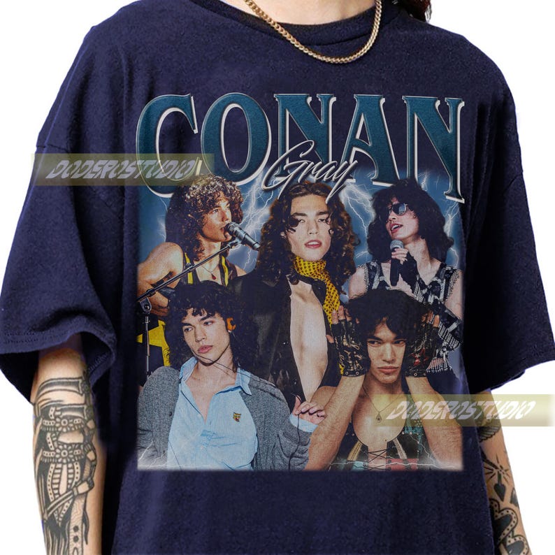 May include: Navy blue t-shirt featuring a vintage-style graphic of Conan Gray and the text "CONAN Gray". The graphic includes a group of people with long hair, microphones, and lightning bolts in the background. The shirt has a crew neck.
