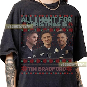 Pode incluir: Camiseta preta com o texto "ALL I WANT FOR CHRISTMAS IS" acima de uma foto de três homens em uniformes policiais. O nome "TIM BRADFORD" está impresso abaixo. O design inclui padrões de flocos de neve e corações vermelhos e verdes.