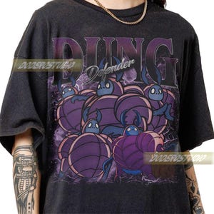 Peut inclure: T-shirt gris foncé avec un motif violet de créatures ressemblant à des scarabées de dessin animé. Le mot "DUNG" est imprimé au-dessus des créatures dans une police violette stylisée, avec "Defender" en dessous. Le design a un aspect vintage et vieilli.