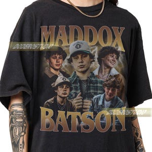 May include: Black t-shirt featuring a collage of images of a person, with the name "MADDOX BATSON" in a gold, vintage-style font. The design includes multiple photos of the person in various poses and outfits.