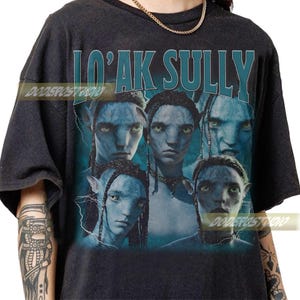 May include: Dark grey t-shirt with a teal graphic of Na'vi characters from the film Avatar, and the text "Lo'ak Sully". The design includes lightning bolt accents and the studio name "DODERPOSTUDIO".