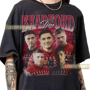 Pode incluir: Camiseta cinza escuro com um design gráfico com a palavra "BRADFORD" em vermelho e o nome "Tim" em branco. O design inclui várias imagens de uma pessoa usando um suéter vermelho com um padrão de cifrão.
