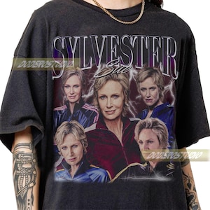 May include: Dark gray t-shirt featuring a graphic of a woman with the name "SYLVESTER" above and "Sue" below. The design includes multiple images of the woman with lightning bolt accents. The shirt has a vintage, distressed look.