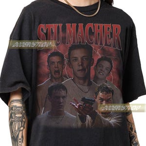 Può includere: T-shirt nera con una grafica di Stu Macher, con più immagini del suo volto e la scritta "STU MACHER" in rosso. Lo sfondo presenta un design a fulmini rossi.