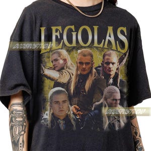 Pu&ograve; includere: T-shirt grigia scura con una grafica di Legolas de Il Signore degli Anelli. Il design include pi&ugrave; immagini del personaggio con lunghi capelli biondi e il nome "LEGOLAS" in lettere dorate.