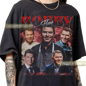 May include: Black t-shirt featuring a graphic of Bobby Flay with multiple images of him, the name "BOBBY" in red, and "Flay" in white script. The design includes red lightning bolt effects. The shirt has a vintage, distressed look.