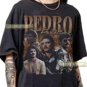 May include: Black t-shirt featuring a graphic of Pedro Pascal with the name "PEDRO Pascal" in a vintage style. The design includes multiple images of the actor. The shirt has a crew neck and short sleeves.