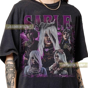 May include: Black t-shirt featuring a graphic design with the text "Sable Ward" in purple. The design includes multiple images of a person with long white hair, with a purple and black background.