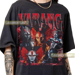 Pode incluir: T-shirt cinzenta escura com um design gráfico. A palavra "VARANG" está em vermelho acima de um grupo de figuras estilizadas com cocares vermelhos e brancos. O design tem um aspeto vintage e desgastado.