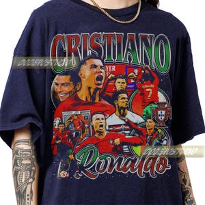 May include: Navy blue t-shirt featuring a collage of images of Cristiano Ronaldo, with the name "Cristiano" in green and red lettering. The word "Ronaldo" is in a cursive font. The shirt has a vintage, distressed look.