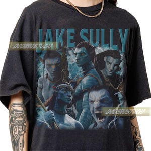 May include: Dark grey t-shirt with a graphic of the character Jake Sully from the film Avatar. The design features multiple images of the character in blue tones, with the name "JAKE SULLY" in blue block letters at the top.