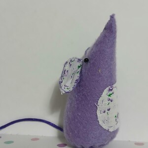 May include: A handcrafted, lavender felt mouse with a pointed head and a long, thin tail. The mouse features a white fabric ear and a white patch with a floral design. A small black bead is used for the eye.