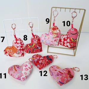May include: Ten heart-shaped keychains made of fabric with various patterns in shades of red, pink, and white. The keychains are hanging on a metal display stand.