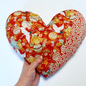 Puede incluir: Almohada roja en forma de corazón con un patrón floral y de grúas. La tela es una mezcla de rojo, blanco y dorado.