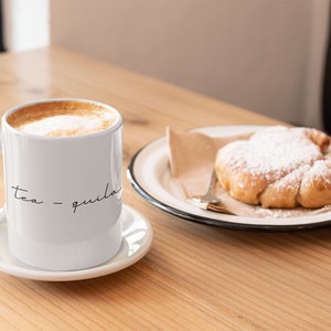 May include: A white ceramic mug with the text "tea -quila" on it, filled with a cappuccino, and a pastry dusted with powdered sugar on a plate.
