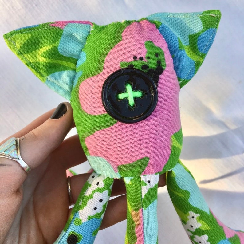 May include: A handmade fabric doll with a whimsical design. The doll features a large black button with a green cross for a face, and is made from colorful floral patterned fabric in pink, green, and blue. It has large ears and long legs.