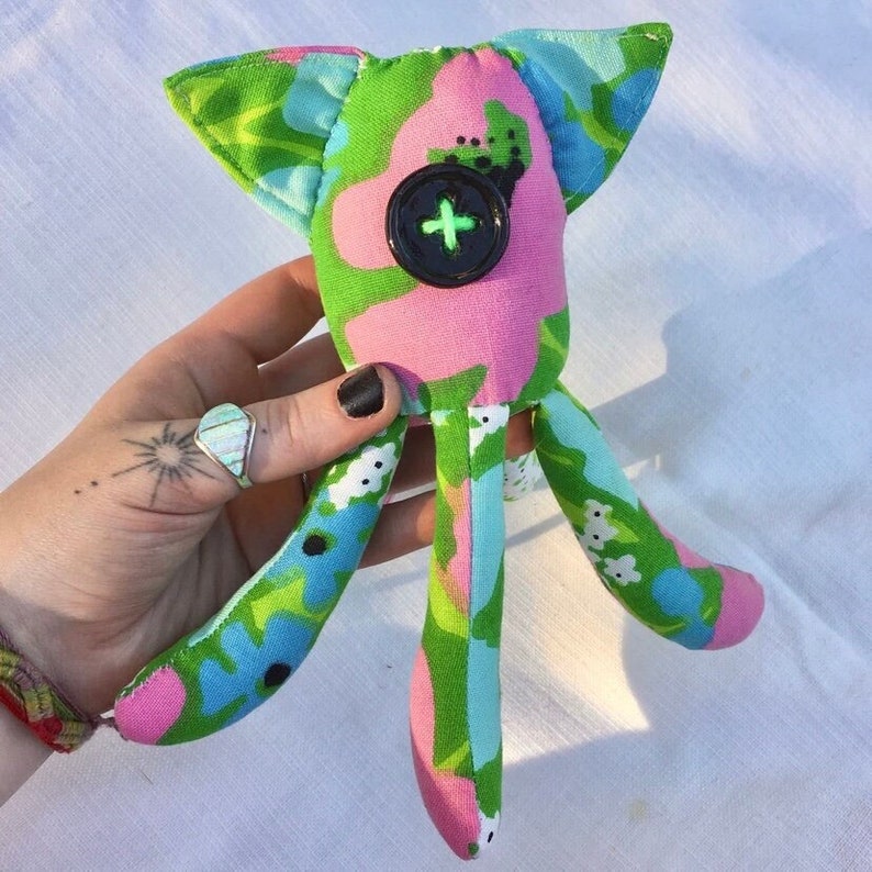 May include: A handmade fabric doll with a whimsical design. The doll features a head with pointed ears, a large black button eye, and four long, slender limbs. The fabric is patterned with pink, green, and blue floral and abstract shapes.