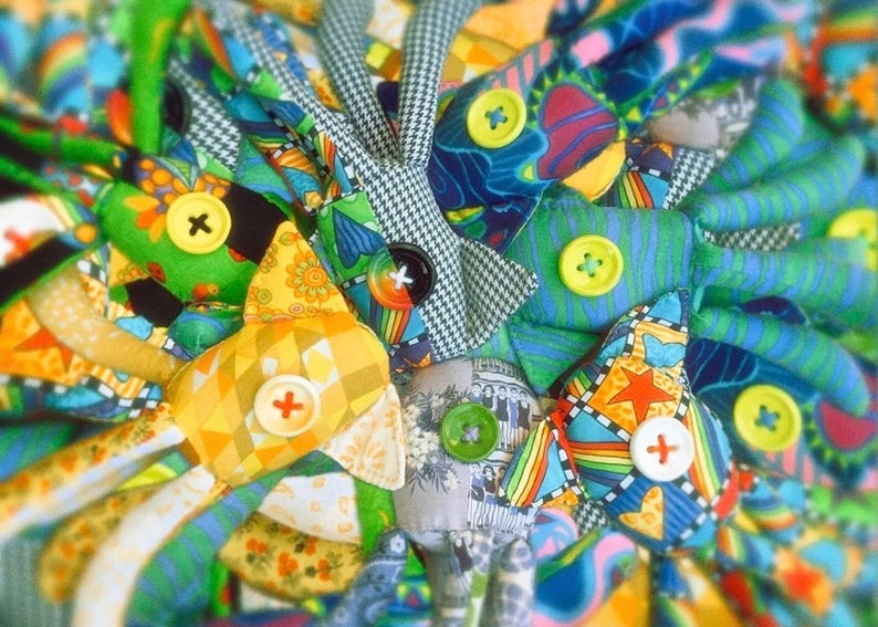 May include: A close-up of a collection of colorful fabric dolls. Each doll features a unique patchwork design with various patterns, including floral, geometric, and houndstooth. Large, decorative buttons in yellow, green, and black secure the pieces.