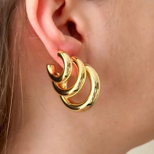 May include: Gold hoop earrings with three interlocking circles. The earrings are large and have a shiny finish.