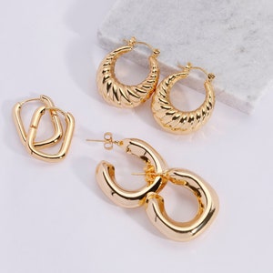 May include: Gold-toned earrings in various styles. The image showcases three pairs: a pair of square hoops, a pair of textured, half-hoop earrings, and a pair of large, rounded hoops. The earrings are displayed on a white surface with a marble accent.