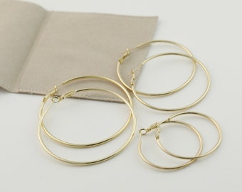 Peut inclure: Plusieurs boucles d'oreilles créoles dorées de différentes tailles. Les boucles d'oreilles sont disposées sur une surface blanche, à côté d'une pochette beige clair. Les créoles varient en taille, de petite à grande, avec un design simple et élégant.