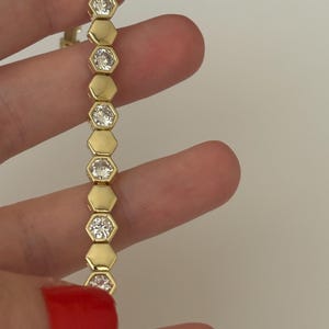 May include: Gold-toned bracelet featuring a series of hexagonal links, each encasing a sparkling, round-cut gemstone. The bracelet is held against a neutral background, showcasing its elegant design and craftsmanship.