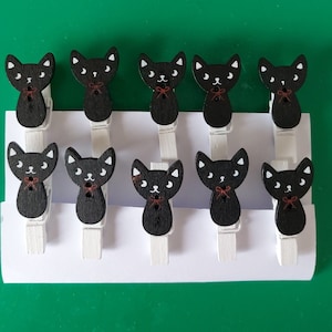Könnte beinhalten: Ein Set mit zehn schwarzen Katzen-Wäscheklammern mit weißen Augen und roten Schleifen. Die Wäscheklammern sind in zwei Reihen zu fünf auf weißem Hintergrund angeordnet.
