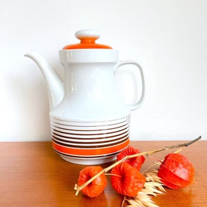 Op de afbeelding: Een witte keramische koffiekan met een bruin en oranje gestreept ontwerp. De kan heeft een deksel met een witte knop en een oranje bovenkant. De kan staat op een houten oppervlak met een takje oranje bessen en gedroogd gras.