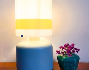 Puede incluir: Una lámpara de mesa con base azul claro mate y pantalla cilíndrica de cristal blanco con una franja amarilla horizontal. Junto a la lámpara hay un pequeño jarrón de cerámica verde oscuro hexagonal con flores moradas.