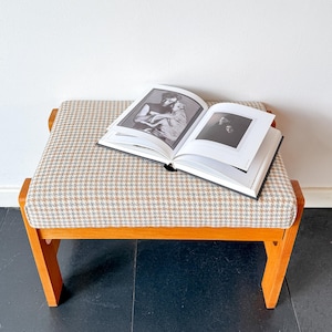 Puede incluir: Un banco de madera con un cojín con estampado de pata de gallo. Un libro abierto con fotografías en blanco y negro descansa sobre el banco. El banco tiene un marco de madera marrón claro y está contra una pared blanca.
