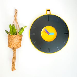 May include: A black and yellow clock with a yellow face and black hands. The clock is hanging on a white wall next to a hanging plant in a woven basket.