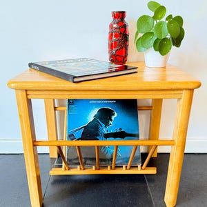 Könnte beinhalten: Ein Beistelltisch aus hellem Holz mit eingebautem Schallplattenhalter. Eine rot-schwarze Vase, eine Topfpflanze und ein Buch stehen darauf. Ein Johnny Cash-Album wird im Schallplattenhalter ausgestellt. Der Tisch ist aus hellem Holz gefertigt.