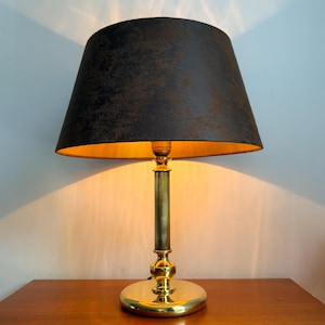 Op de afbeelding: Een tafellamp met een gepolijste goudkleurige metalen voet, een geribbelde cilindrische stang en een ronde voet. De lamp is voorzien van een brede, taps toelopende trommelkap in een donkere, getextureerde antracietgrijze afwerking met een gouden voering.